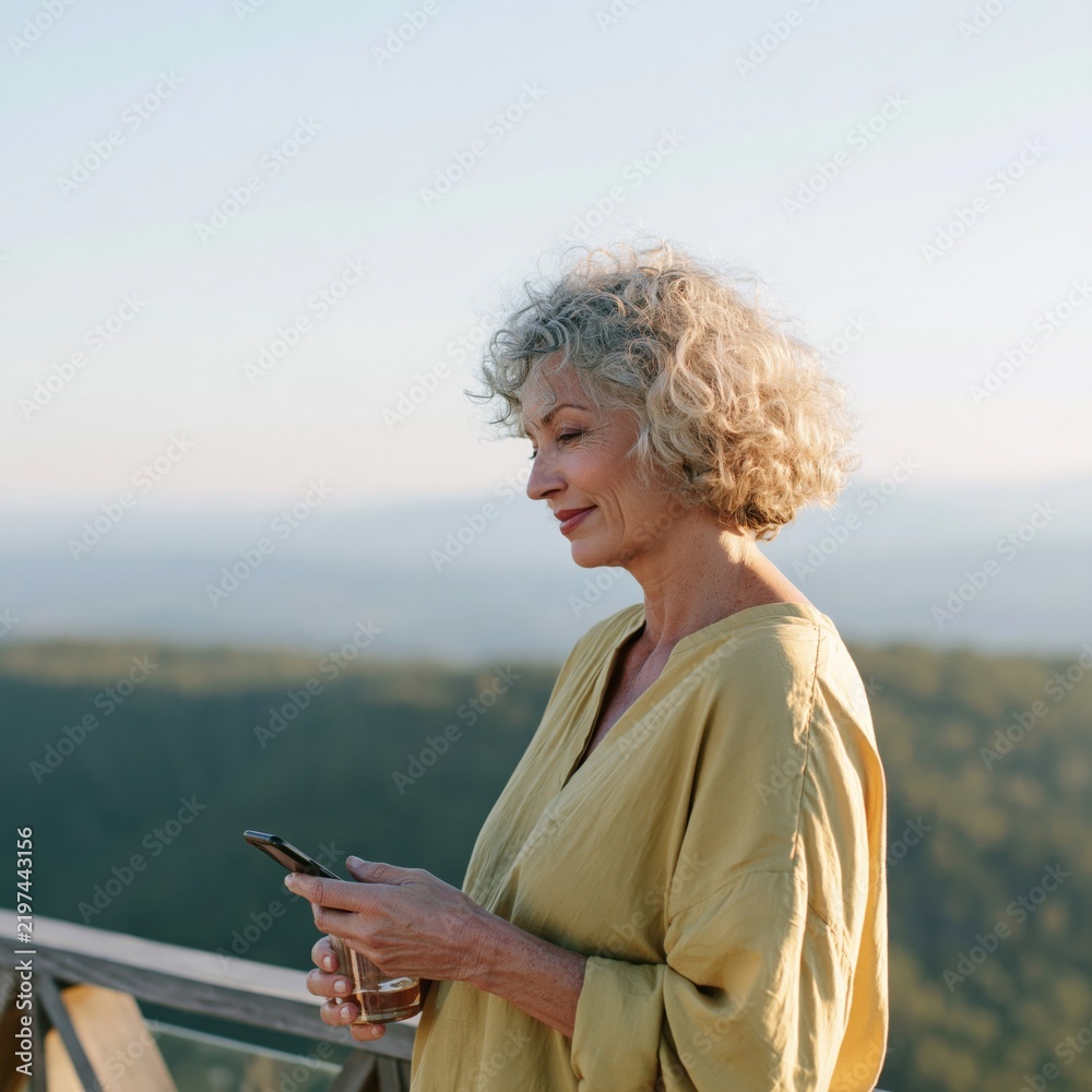 Fototapeta premium Senior woman using a smartphone on a wooden balcony with panoramic valley view for wellness lifestyle background and retreat design