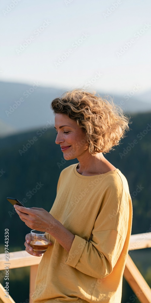 Fototapeta premium Middle aged woman with curly hair in a yellow sweater checking phone on a mountain balcony holding a glass — lifestyle outdoor portrait background for banner and wallpaper