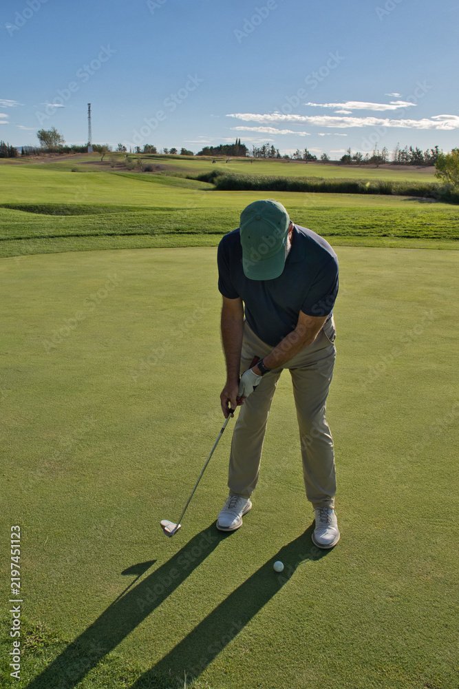 Obraz premium Senior golfer standing over the ball ready to putt, focused expression, green course stretching behind him.