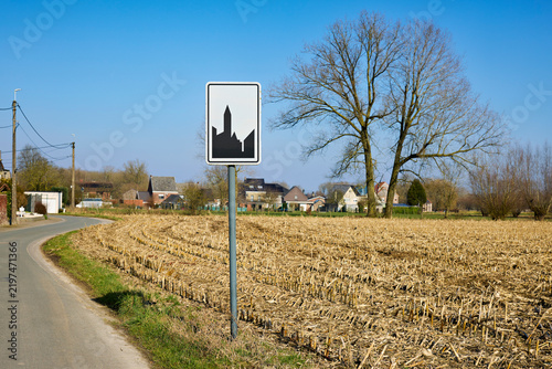 Belgian road sign for a built-up area along a rural asphalt road, with a harvested field, village houses, bare trees, and a clear blue sky in the background.
