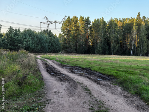 Muddy dirt road leading to forest with power line tower under clear sky
