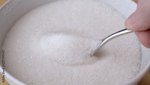 Konstfotografi Close-up view of a white ceramic bowl filled with granulated white sugar, featuring a metal spoon scooping the sugar crystals by a hand