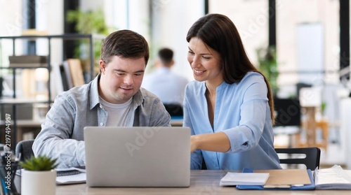 Friendly female mentor in soft blue blouse guiding young male coworker with Down syndrome at laptop in bright modern office, inclusive workplace collaboration