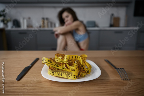 Depressed anorexic woman looking at plate with measuring tape served for dinner