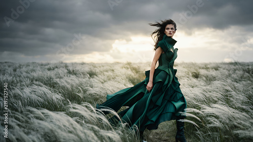Young woman in a flowing dark green dress walking through a windswept field under a stormy sky