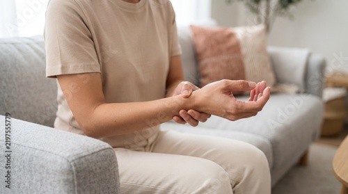 Woman sitting on a comfortable gray sofa wearing a neutral beige t-shirt while gently holding her sore wrist to alleviate sharp pain and physical discomfort