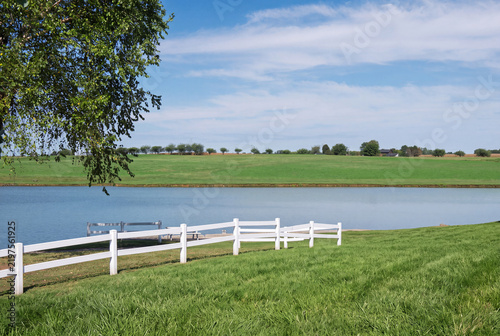 White fence borders calm water. Lush green grass covers the foreground.