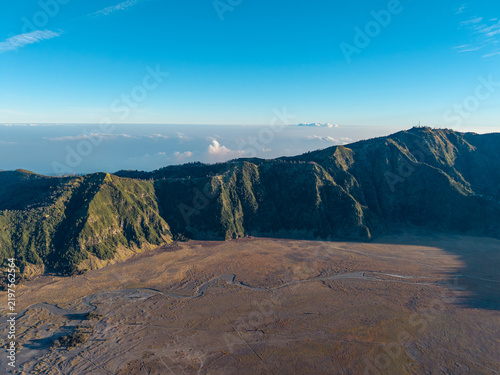 Morning drone panorama reveals Mount Bromo rugged volcanic caldera, forested ridges, ash plains, clouds, and distant mountains.