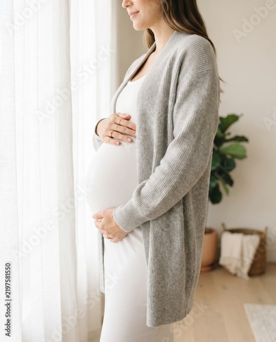 A pregnant woman is standing beside a window in a bright room, wearing a gray sweater over a white dress while gently holding her belly.