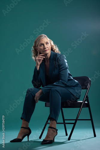 Senior woman sitting on black metal folding chair in studio wearing teal pantsuit and heels