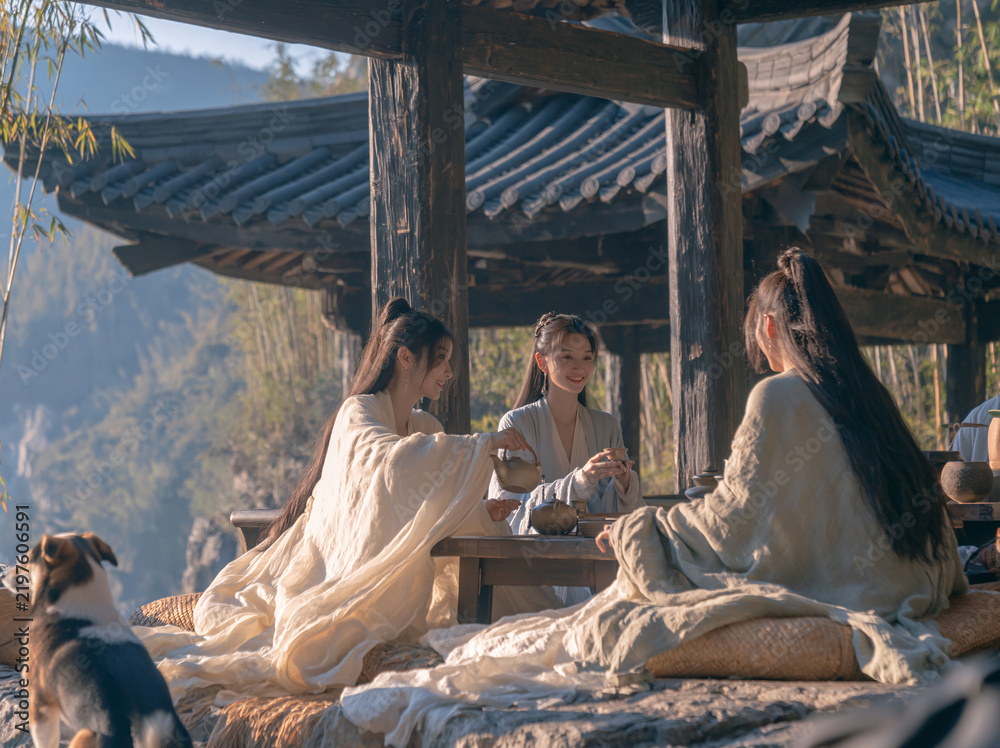 Obraz premium Three people in flowing ancient hanfu share tea beneath a weathered wooden pavilion, mountains and a small dog in the background.