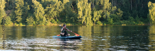 Cuadro en lienzo A young Caucasian man paddles on a blue paddle board in a calm lake surrounded by lush green trees