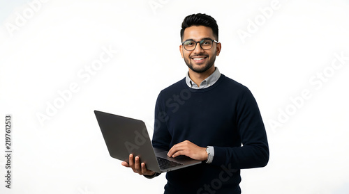 A smiling Indian man wearing glasses holds a laptop computer and stands against a white background.