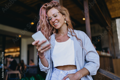 Caucasian female student in glasses smiling warmly while texting on smartphone, receiving positive online exam result notification standing in modern coworking studio workspace.