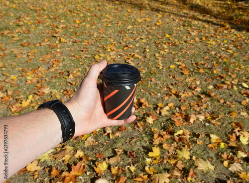 A cup of hot tea or coffee. A drink held in a man's hand. Fallen autumn leaves lie on the ground around a cup of hot coffee or tea. A warming drink on a clear autumn day.