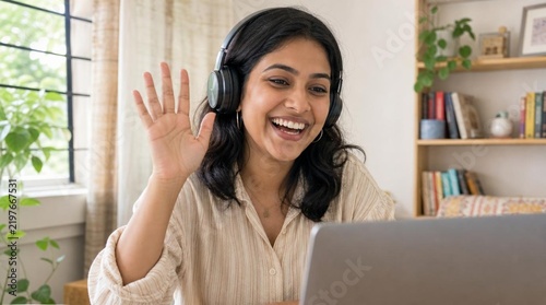 Cheerful young Indian woman wearing headphones, waving hello and laughing warmly during a video call on laptop webcam in bright cozy home office setting with bookshelf and plants