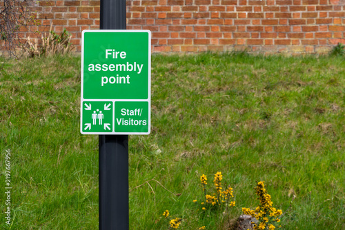 Obraz na plátně Green fire assembly point sign on black post, grassy bank