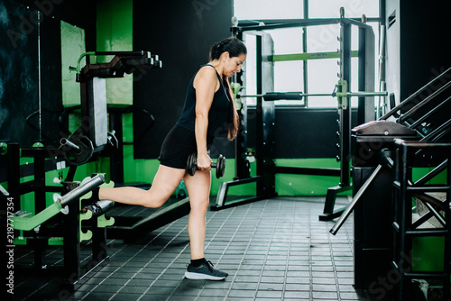 Fotografie A woman performs a dumbbell exercise in a modern gym, leaning forward with controlled posture while developing lower-body strength and overall physical conditioning