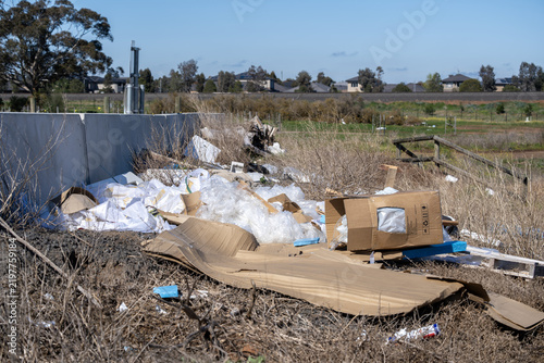 Illegally dumped waste, cardboard boxes, plastic packaging, and debris on a vacant peri-urban road reserve bordering new residential housing estates. Truganina, Melbourne, Victoria, Australia
