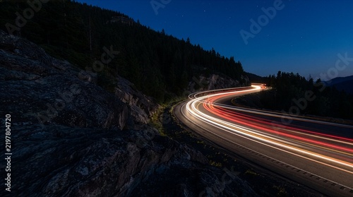 Long exposure light trails curve along winding road at night, illustrating transportation, speed, motion, travel, traffic flow and dynamic highway movement through scenic landscape.