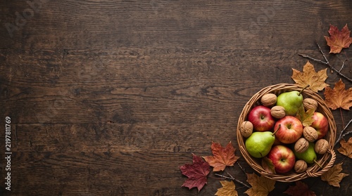 Rustic autumn harvest background with apples and pears, wicker basket of seasonal fruit on dark wood, fall foliage and walnuts on wooden table