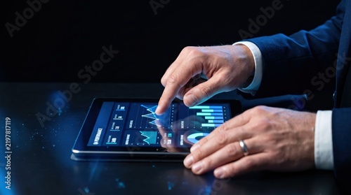 Close up of businessman hands using tablet computer to analyze financial performance charts and graphs in dark office, mobile business analytics and data concept