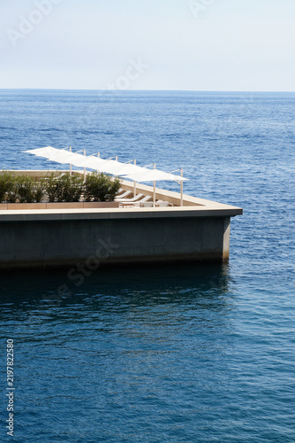 Konstfotografi Modern seaside deck with a row of white sun loungers and umbrellas overlooking a vast blue sea under a pale, cloudless sky