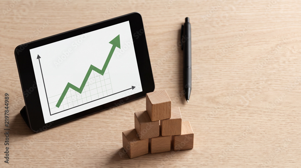 Obraz premium Business Growth and Financial Success Concept with Digital Tablet Showing Rising Green Stock Market Graph Next to Pyramid of Wooden Blocks and a Professional Pen on a Light Wood Desk Surface