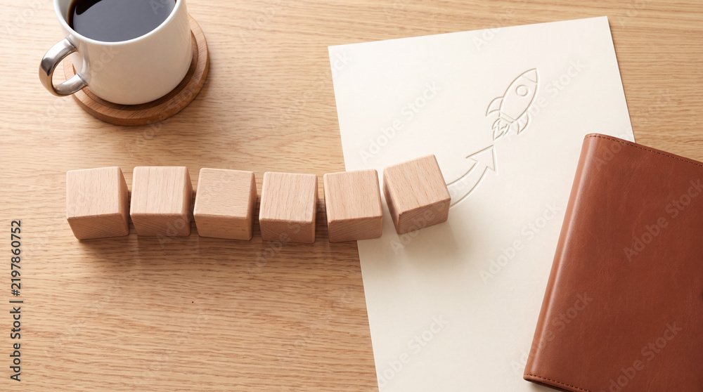 Obraz premium Conceptual business growth image with wooden blocks leading to a rocket launch symbol on paper with a cup of coffee and leather notebook on a desk, symbolizing startup success and planning.