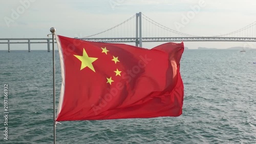 Chinese national flag waving in the wind with the Xinghai Bridge visible in the background in Dalian, Liaoning, China.