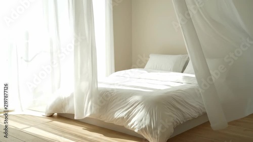 A sunlit bedroom with white curtains blowing gently while sunlight streams through the window onto the bed