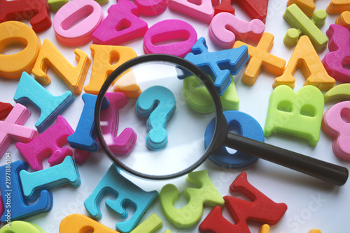 Magnifying glass focuses on a bright blue question mark among scattered colorful foam alphabet letters, suggesting learning, curiosity, education, children's play, investigation and problem solving.