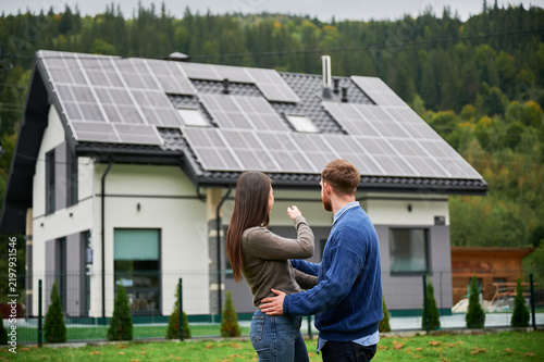 Couple looking at house with solar panels, woman pointing at rooftop system. Homeowners planning renewable energy use, eco friendly house and sustainable lifestyle concept