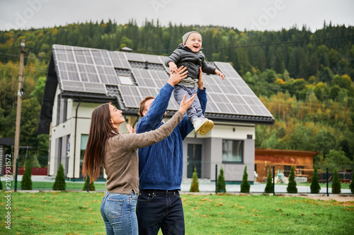 Parents lifting happy child in air on lawn near modern house with solar panels. Photovoltaic rooftop system, renewable energy concept, eco friendly family lifestyle outdoors