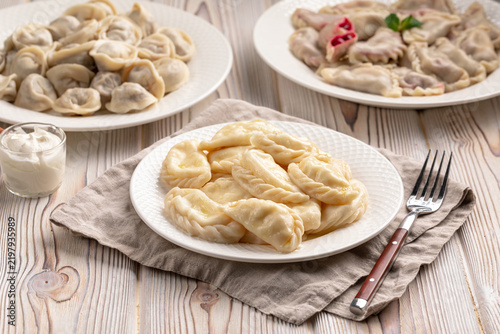 Three white plates with assorted cooked vareniki with different fillings and meat dumplings on rustic wooden background