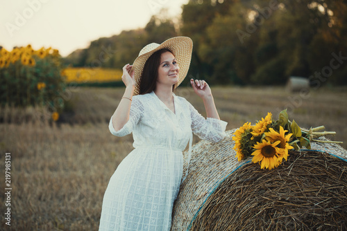 woman in a white dress holding a bouquet of wild flowers with sunflowers in a mowed field with straw bales