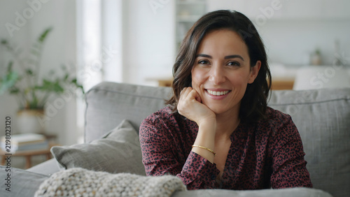 A smiling young woman with dark hair sitting on a comfortable grey sofa in a bright living room, resting her chin on her hand and looking at camera
