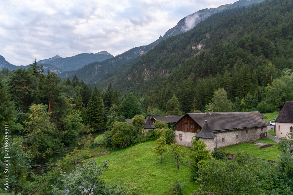 Obraz premium Traditional stone and timber buildings stand in a lush valley near Fernsteinsee in Tyrol, Austria. Forested Alpine slopes, meadow and low mountain clouds create a scenic rural setting for European tra