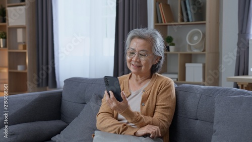 Картината върху платно Happy senior Asian woman looks at a smartphone while relaxing on a sofa at home, showing positive aging, independence, digital connection, mobile technology and a comfortable retirement lifestyle