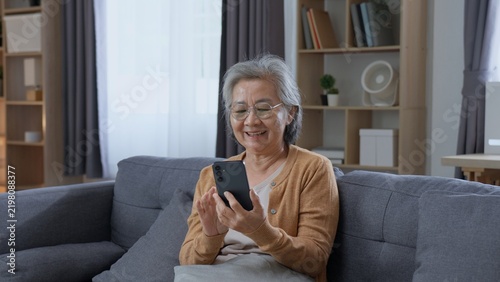 Фотография Happy senior Asian woman uses a smartphone while sitting on a sofa at home, showing digital technology, online communication, active aging, independence and a positive retirement lifestyle