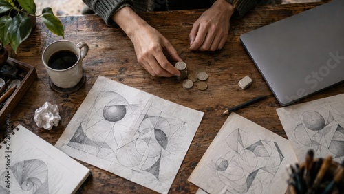 Person stacking coins on wooden desk with sketches and laptop