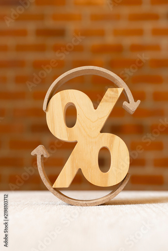 Loan refinancing and interest rate cycle concept, wooden percentage sign encircled by curved arrows on table against brick wall, symbolizing debt restructuring, renewal, and turnover.