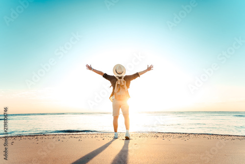 Happy man with backpack standing with arms up at the beach - Delightful tourist enjoying summer vacation by the seaside - Traveling life style and well being concept