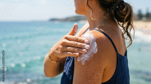 A woman applying sunscreen to her shoulder, with the ocean in the background.