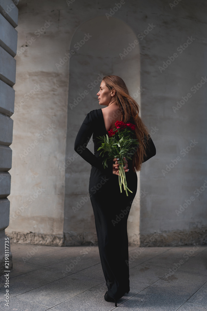 Fototapeta premium Red Paeonia lactiflora flowers held by a woman in a black dress standing within an arched stone corridor