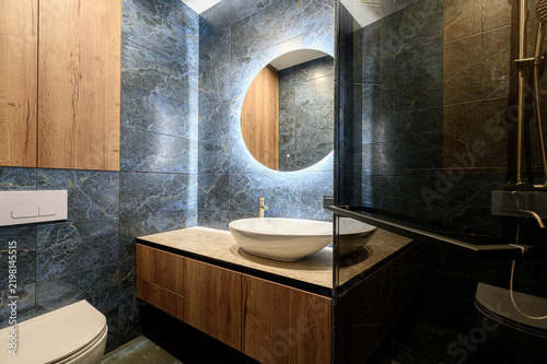 Compact bathroom features dark marble tiles, wooden vanity, vessel sink, round illuminated mirror, concealed toilet, glass partition, brass shower fittings, sleek modern design