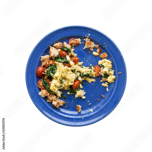 Unfinished scrambled eggs and toast crusts on a blue plate isolated. Partially eaten breakfast leftovers with spinach and cherry tomatoes. Top view of messy remaining morning meal scraps.