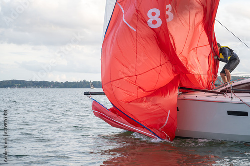 Segler birgt den leuchtend roten Gennaker einer Yacht auf leicht welligem Meer vor bewölktem Himmel und grüner Küstenlandschaft.