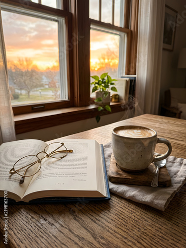 Cappuccino with latte art beside open book and glasses on oak table at sundown.