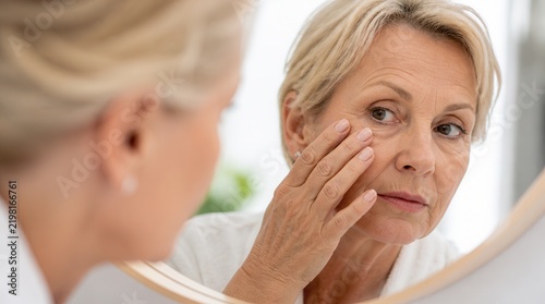 Mature woman applying eye cream or skincare treatment while looking at mirror in a bright, modern bathroom